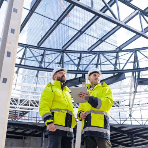 Cert IV Training and Assessment. Two men wearing yellow Hi-Vis and white hardhats holding a electronic tablet standing under large construction.