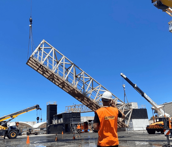 Rigging Intermediate and Advanced. A rigger in an orange high-visibility vest and white helmet communicating via radio on a construction site with cranes in the background