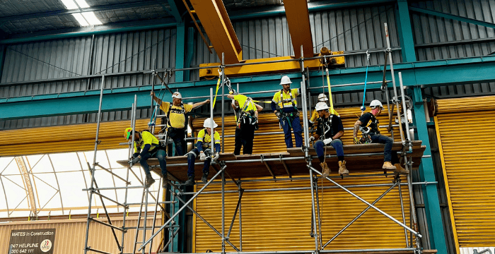 Workers in safety harnesses and helmets working on an industrial scaffolding structure inside a facility, demonstrating the techniques and skills taught in the Scaffolding Intermediate course